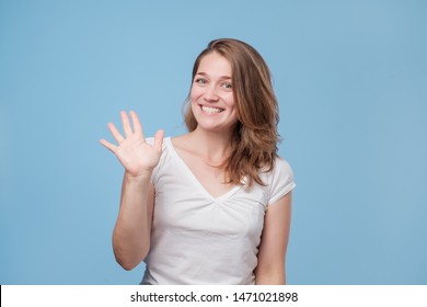 Attractive friendly looking young woman smiling happily, saying Hello, Hi or Bye, waving hand. Showing five fingers. Studio shot
