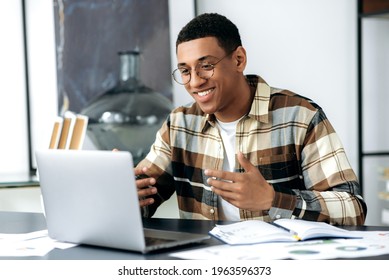 Attractive Friendly Mixed Race Latino Freelancer Guy With Glasses, Sits At A Desk, Uses Laptop, Have A Video Call, Communicate With Colleagues Or Friends By Video Conference, Gesturing Hands, Smiling