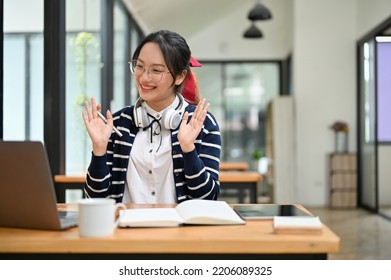 Attractive And Friendly Young Asian Female Student Uses A Laptop To Study An Online Lesson, Waving Hands To Her Friends, Say Hi Or Greeting. 