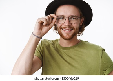 Attractive Friendly-looking Charismatic Young Bearded Guy In Glasses And Hat Touching Frames Of Eyewear And Smiling Broadly At Camera With Pleased Carefree Expression Over Gray Background