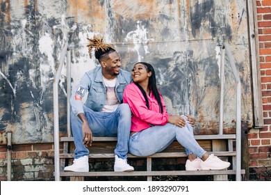 An Attractive And Fun African-American Couple Sitting Outside On Steps In An Urban Setting With A Jacket And Denim Jeans Laughing Together And Having Fun