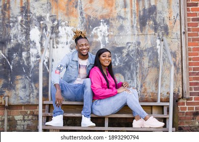 An Attractive And Fun African-American Couple Sitting Outside On Steps In An Urban Setting With A Jacket And Denim Jeans Laughing Together And Having Fun