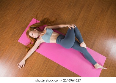 Attractive ginger girl practicing yoga in Reclined Spinal Twist pose. Woman stretching on pink mat in Supta Matsyendrasana yoga pose. Fitness training at home or gym class. Healthy lifestyle concept.