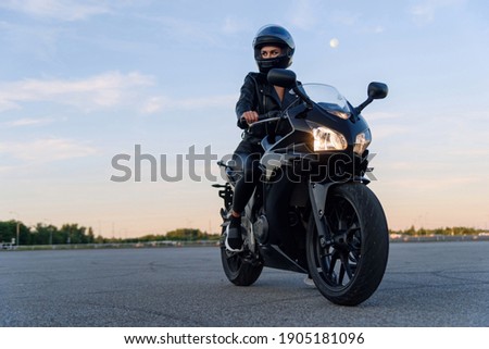 Attractive girl in black leather jacket, pants and helmet on outdoors parking rides on stylish sports motorcycle at sunset.