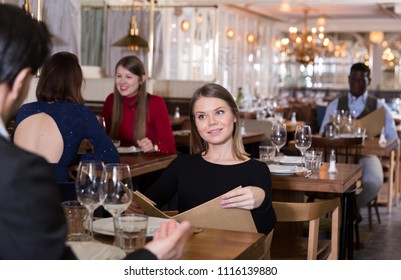 Attractive girl with boyfriend choosing meal from menu in cozy restaurant