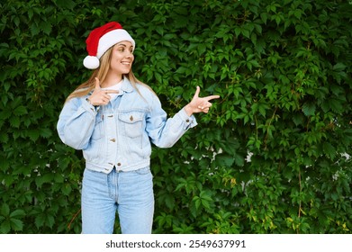 Attractive girl in a Christmas cap points with a fingers green background. Young European woman wearing Santa Claus hat points index fingers to center of green background. Christmas. New Year. 2025 - Powered by Shutterstock