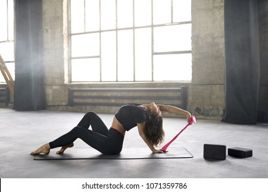 Attractive girl with closed eyes in a black sportswear engaged in yoga on the windows background in a loft style hall. She sits on the mat and does a backward deflection and pulls a pink stretch band. - Powered by Shutterstock