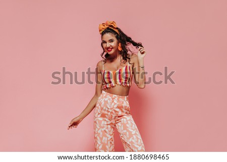Attractive girl with cool hairstyle in orange earrings and fashionable bright clothes smiling and looking into camera on pink backdrop..