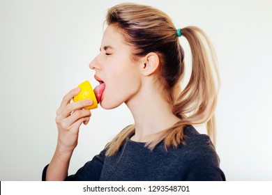 An attractive girl holds a lemon in her hand and licks it with a squint of sour taste.