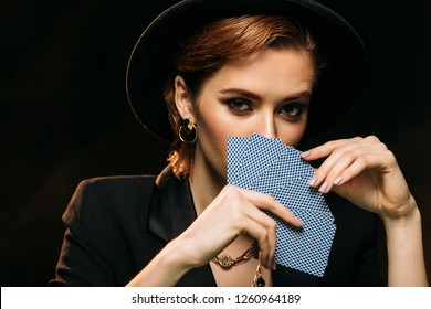 attractive girl in jacket and hat covering face with poker cards isolated on black, looking at camera