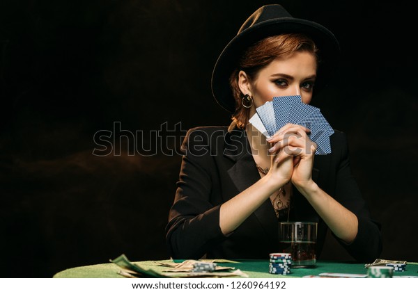 attractive girl in jacket and hat\
covering face with poker cards and looking at camera in\
casino