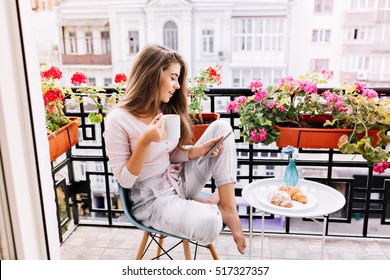 Attractive girl with long hair in pajama having breakfast on balcony in the morning in city. She holds a cup, reading on tablet