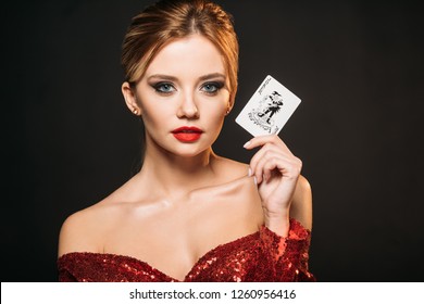 attractive girl in red shiny dress holding joker card and looking at camera isolated on black