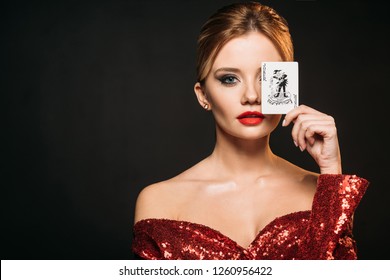 attractive girl in red shiny dress covering eye with joker card isolated on black