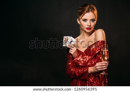 attractive girl in red shiny dress holding joker and queen of hearts cards isolated on black, looking at camera