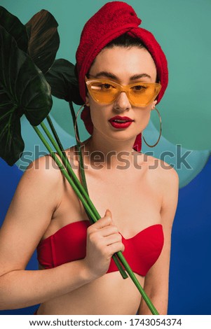 attractive girl in red towel swimsuit and sunglasses holding tropical palm leaves on blue and turquoise