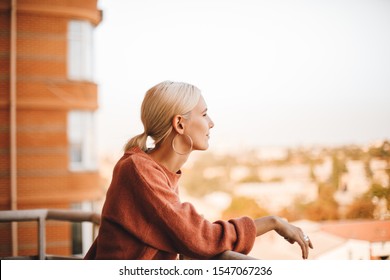 Attractive girl with short blonde hair in pink sweater on balcony in the morning in city. Adorable sensual girl laughing at balcony. Photo of cheerful caucasian woman with beautiful smile.