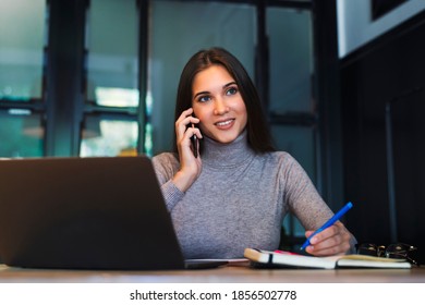 Attraktives Mädchen sitzt am Tisch vor dem Laptop und spricht auf dem Handy, Noten in Notebook. 