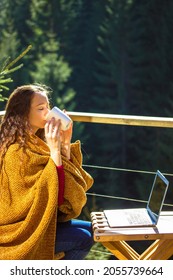 Attractive girl sitting on balcony terrace wrapped in yellow plaid, drinking tea or coffee from white cup. Brunette woman works remotely on laptop, online. House in pine wood at autumn day. Cozy place