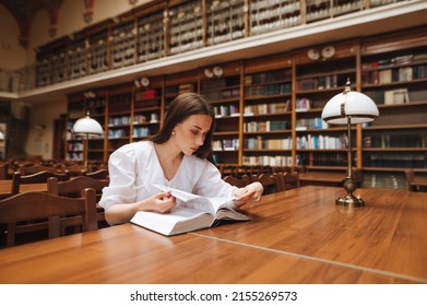 Attractive girl student studying at the university campus library, reading a book sitting at a table with a serious face. - Shutterstock ID 2155269573