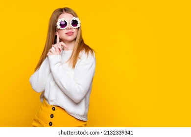 Attractive girl wears white sweater and stylish daisies sunglasses keeps hands together looks away with dreamy expression dreams. Young woman wait oncoming of spring on yellow background. Lifestyle