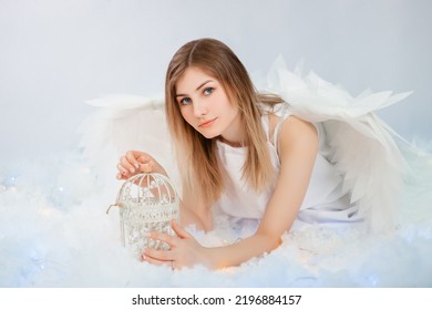 An attractive girl in a white tunic with large white wings behind her back poses while lying in white luminous clouds.