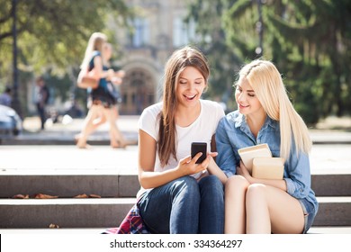 Attractive Girls Are Sitting On Steps. They Are Looking At The Mobile Phone With Joy And Smiling. The Blond Student Is Holding Books. Copy Space In Left Side