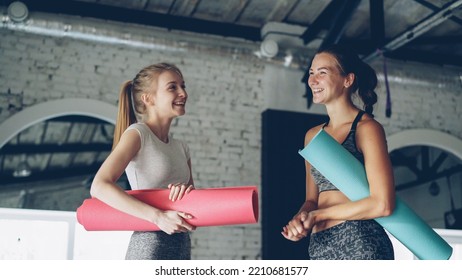 Attractive Girls Are Talking And Laughing In Large Modern Sports Center. Women Are Holding Colorful Yoga Mats And Wearing Fashionable Sports Clothing. Fun In Yoga Studio Concept.