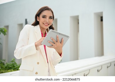 Attractive And Gorgeous Asian Female CEO Or Businesswoman In White Suit, Holding A Digital Tablet, Standing Outside Of The Building, Looking And Smiling At The Camera.