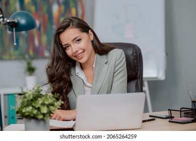 An Attractive Gorgeous Woman With Long Brown Hair And Eyes Is Sitting In Front Of Computer In Corporation, Working Behind Desk In An Office, Looking At Camera With Smile, Calendar Lying Under Hand