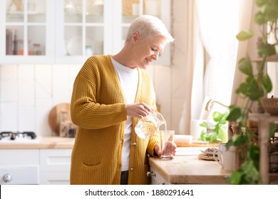 Attractive grandmother in casual outfit pouring spring water into glass in kitchen, copy space. Side view of grey-haired senior woman drinking water while cooking, home interior - Powered by Shutterstock