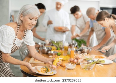 Attractive gray haired elderly woman wearing striped apron honing culinary skills during group cooking classes, enthusiastically garnishing and seasoning with sauce finished dish before serving - Powered by Shutterstock