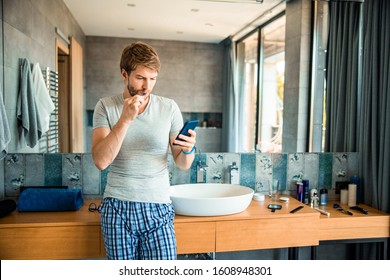 Attractive guy cleaning teeth with toothbrush and checking messages on smartphone stock photo - Powered by Shutterstock