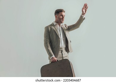 Attractive Guy With Open Collar Shirt, Holding Luggage, Looking To Side And Holding Hand Up Getting A Cab To Airport, Posing In Front Of Beige Background In Studio