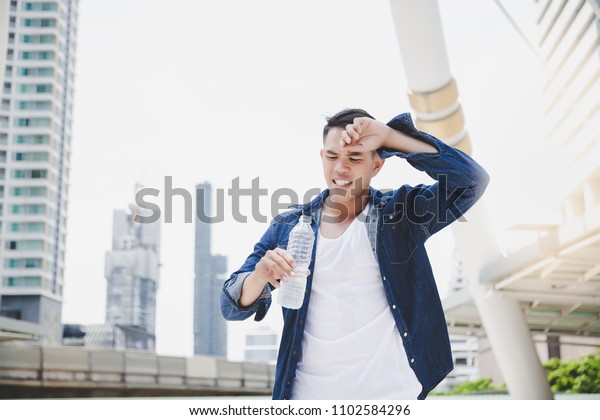 Attractive handsome asian man gets thirsty\
because of hot weather in the summer season. Cool guy feels\
exhausting and holds the bottle of cold water. He wears denim\
jacket. copy space, city\
background