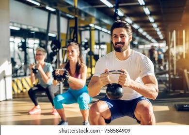 Attractive Handsome Bearded Man Holding Kettlebell And Doing Squats In A Fitness Group With Two Girls In The Modern Gym.