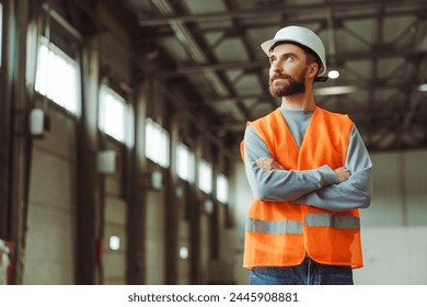 Attractive, handsome factory man wearing hard hat and workwear with crossed arms standing in warehouse, looking away at copy space. Concept of advertisement, engineer job - Powered by Shutterstock