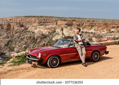 An Attractive Handsome Man Mate Is Leaning On A Retro Car In A Classic Suit On A Sunny Afternoon On A Sandy Desert Road