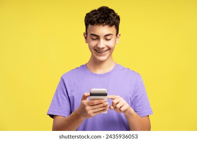 Attractive handsome teenager boy with dental braces holding mobile phone, using mobile app, ordering, text message, watching video isolated on yellow background. Technology concept, online shopping - Powered by Shutterstock