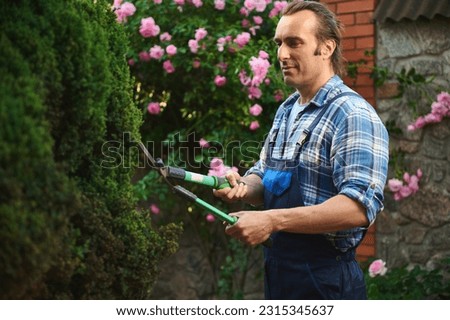 Attractive handsome young adult Latin American professional male gardener, landscaper, horticulturist cutting plants, using pruning shears for trimming and tending hedges in the garden or backyard