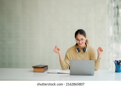 Attractive and happy Asian female student wearing headphones on the neck sitting at the living room table smiling and at work on laptop, notebook, young entrepreneur or freelance business idea. - Shutterstock ID 2162865807