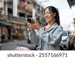 An attractive, happy Asian female tourist in a denim jacket enjoying taking photos with her camera while exploring the city, walking down the street. people and lifestyle concepts