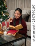 An attractive, happy Asian woman in a cozy red sweater sits in her living room, enjoying a book and some snacks during the Christmas holiday, spending the festive season at home alone.