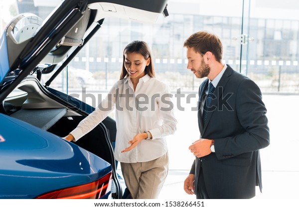 attractive and happy car dealer gesturing near blue car\
and bearded man 
