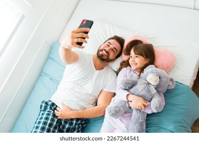 Attractive happy dad taking a selfie with a smartphone relaxing in pajamas in bed with her young child - Powered by Shutterstock