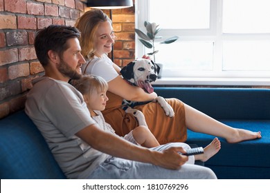 Attractive Happy Family With Dog Watch Tv In Living Room, Young Man, Woman, Daughter And Pet Dog Sit On Sofa, Weekends