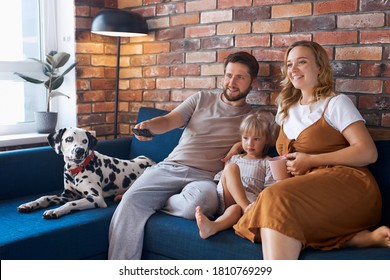 Attractive Happy Family With Dog Watch Tv In Living Room, Young Man, Woman, Daughter And Pet Dog Sit On Sofa, Weekends