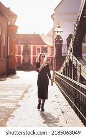 Attractive Happy Girl In A Black Dress  Walks Along A City Street With A Glass Of Wine. Glamorous Lifestyle. Fashion.