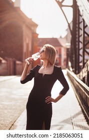 Attractive Happy Girl In A Black Dress  Walks Along A City Street With A Glass Of Wine. Glamorous Lifestyle. Fashion.