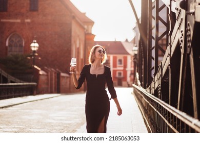 Attractive Happy Girl In A Black Dress  Walks Along A City Street With A Glass Of Wine. Glamorous Lifestyle. Fashion.